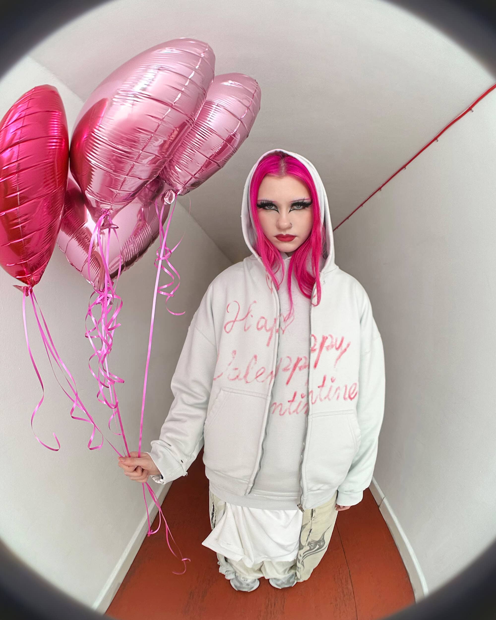 A young woman with bright pink hair and a white hoodie holds a bunch of shiny pink heart-shaped balloons in a narrow hallway.