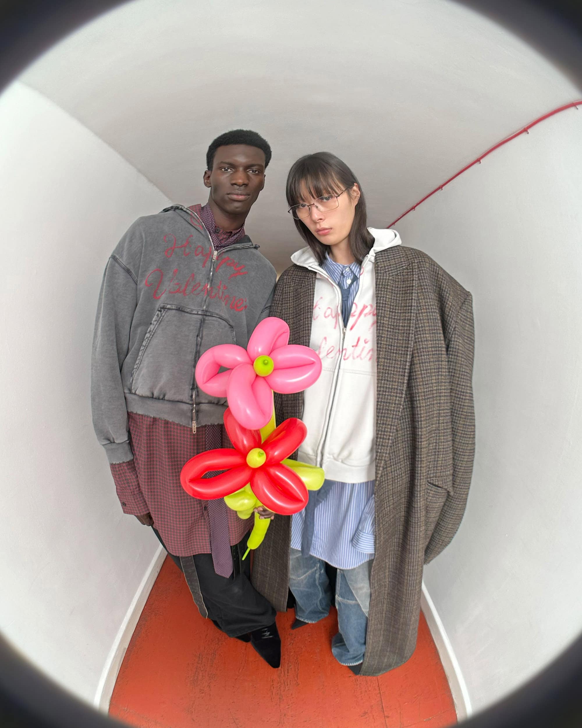 Two models holding bright pink and red balloon flowers inside a curving white tunnel for Balenciaga.
