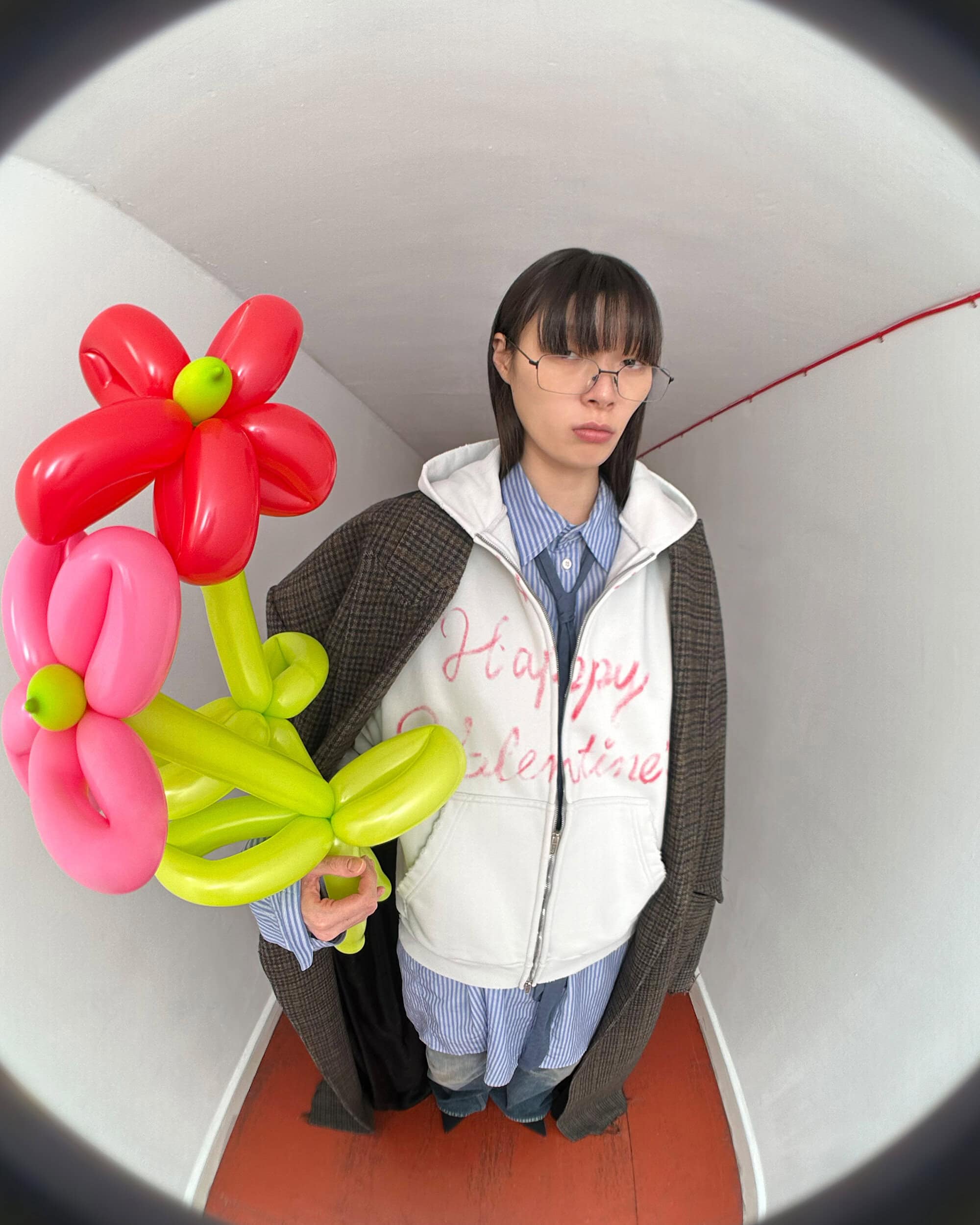 Model holding twisted balloon flowers wearing a white hoodie and plaid coat in an interior hallway