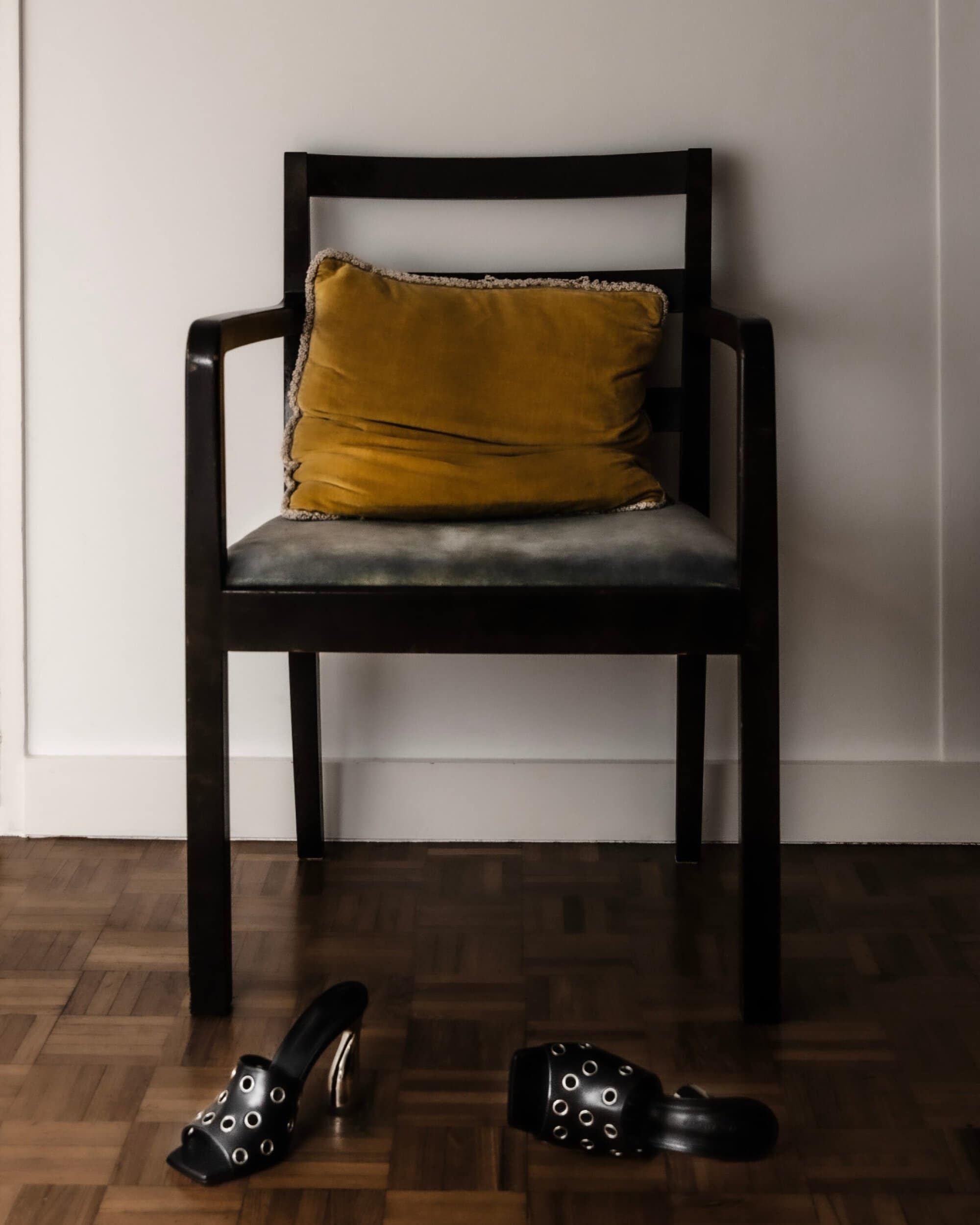 Black wooden armchair with yellow velvet pillow and black high heel mules on parquet floor against white wall
