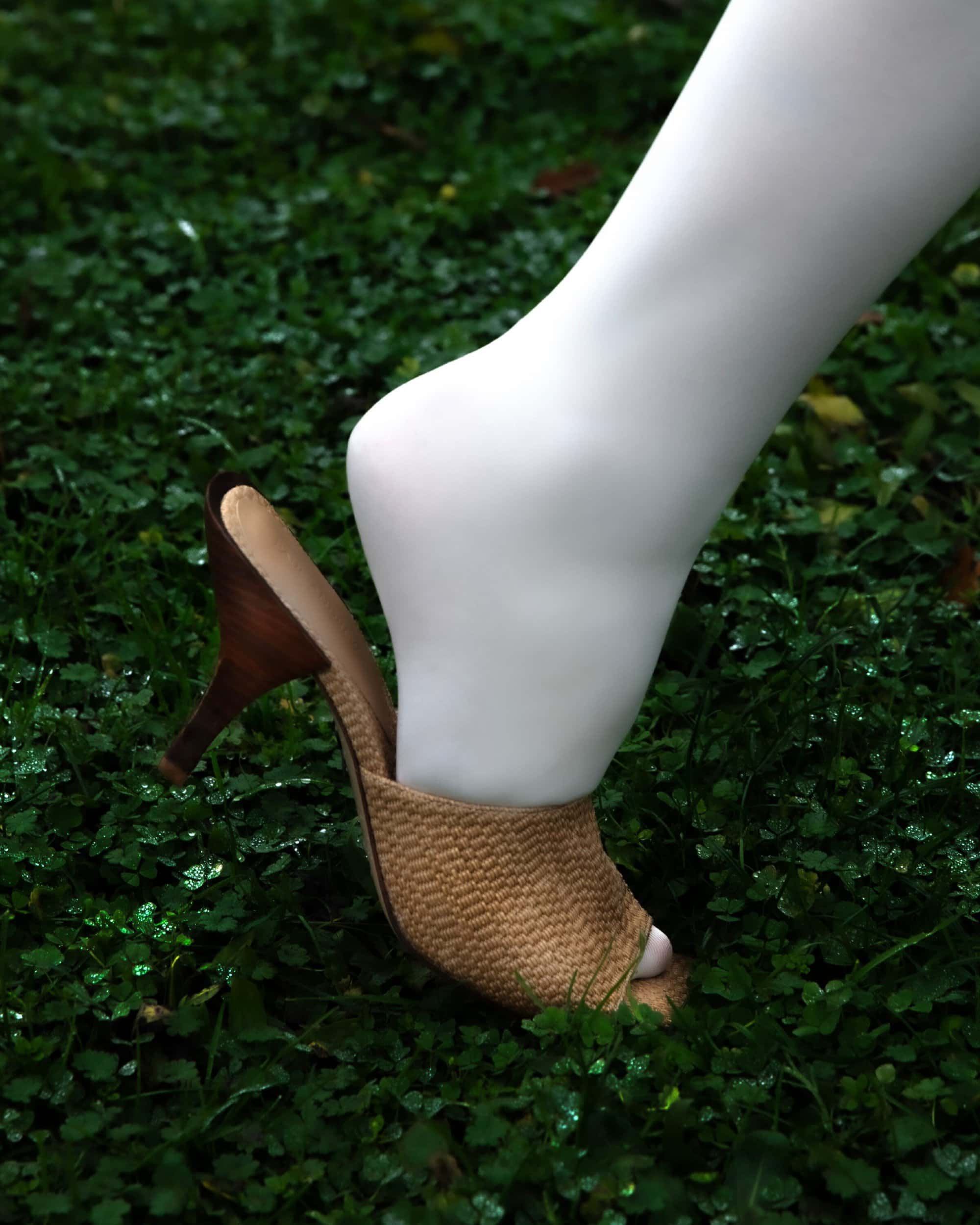 Close-up shot of a white-hosed leg wearing a high-heeled mule standing on green grass for Paul Andrew