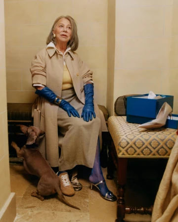 Elderly woman seated wearing a beige trench coat and blue leather gloves alongside a dachshund in an interior space