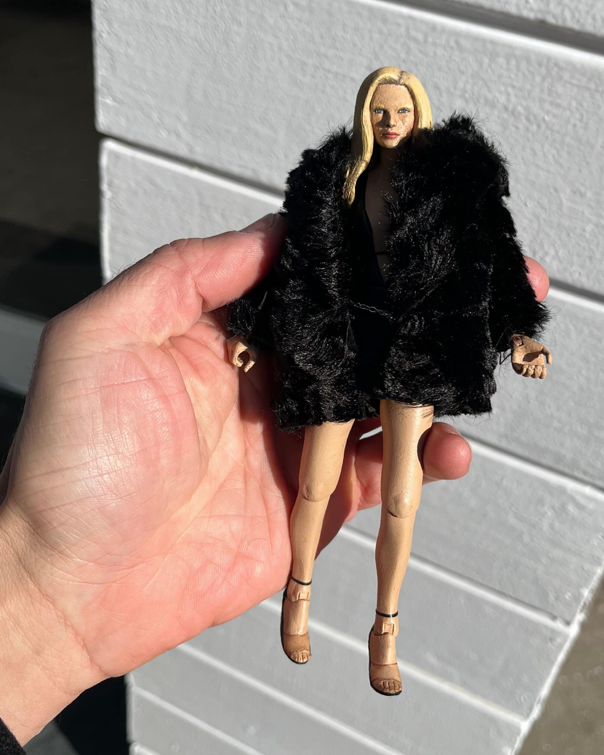 Hand holding a miniature fashion doll wearing a black faux-fur coat and heels against a white panelled wall.