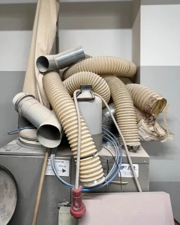Industrial workshop corner featuring a tangle of ribbed beige vacuum hoses and metal ducting pipes stacked on grey machinery