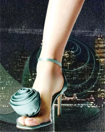 Close-up of a leg wearing a sage green satin high heel sandal with a large floral embellishment against a textured night cityscape background.
