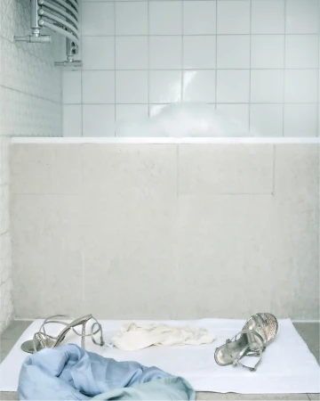 Silver strappy sandals and discarded clothing resting on a white mat in front of a tiled bathtub