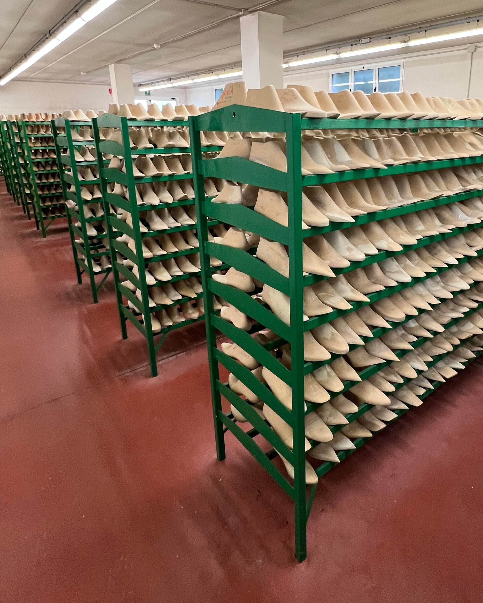 Rows of green metal shelving holding numerous wooden shoe lasts inside a brightly lit manufacturing facility.