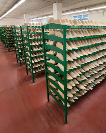 Rows of green metal shelving holding numerous wooden shoe lasts inside a brightly lit manufacturing facility.
