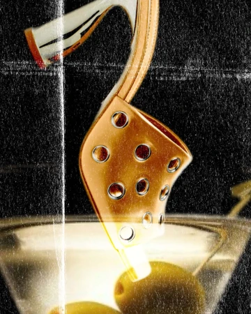 Close-up of a gold high-heeled shoe dipping into a martini glass with olives against a textured dark background.