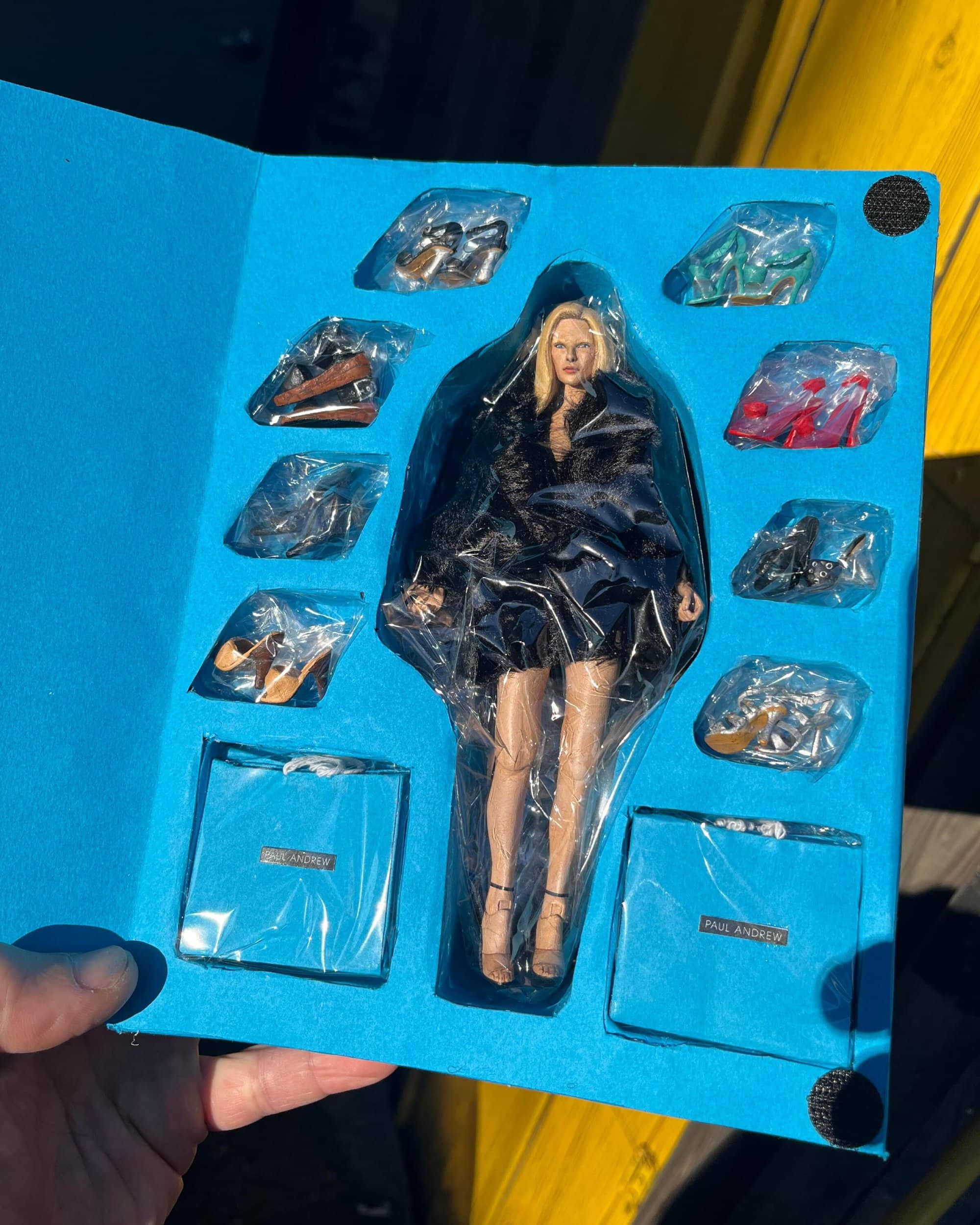 Doll figure in plastic packaging with multiple accessory shoes and bags inside a blue folder for Paul Andrew
