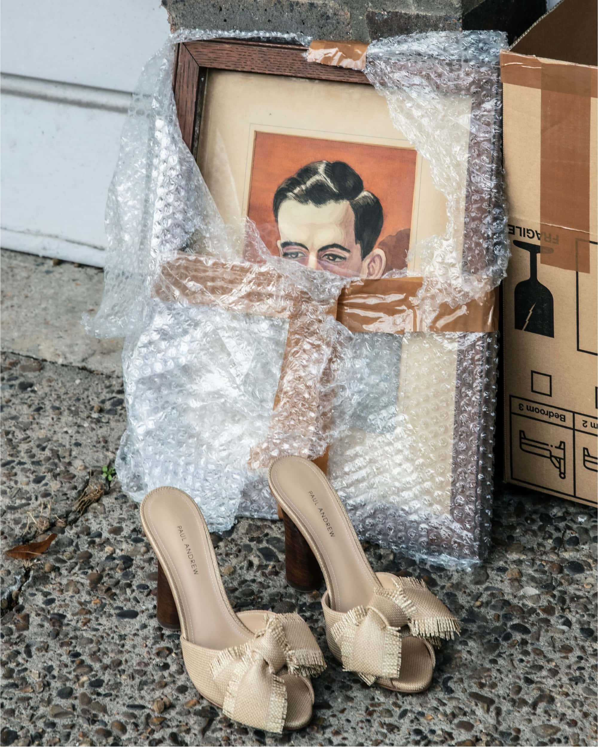A pair of beige heeled sandals with fringed bows rests on stone ground next to a framed portrait wrapped in bubble wrap and a cardboard box.