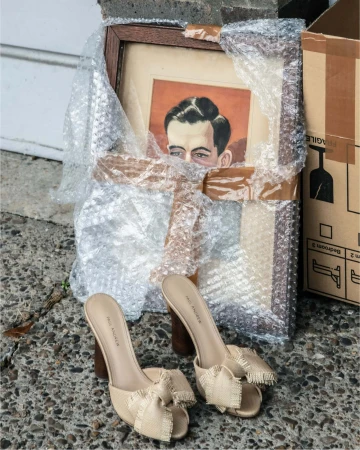A pair of beige heeled sandals with fringed bows rests on stone ground next to a framed portrait wrapped in bubble wrap and a cardboard box.