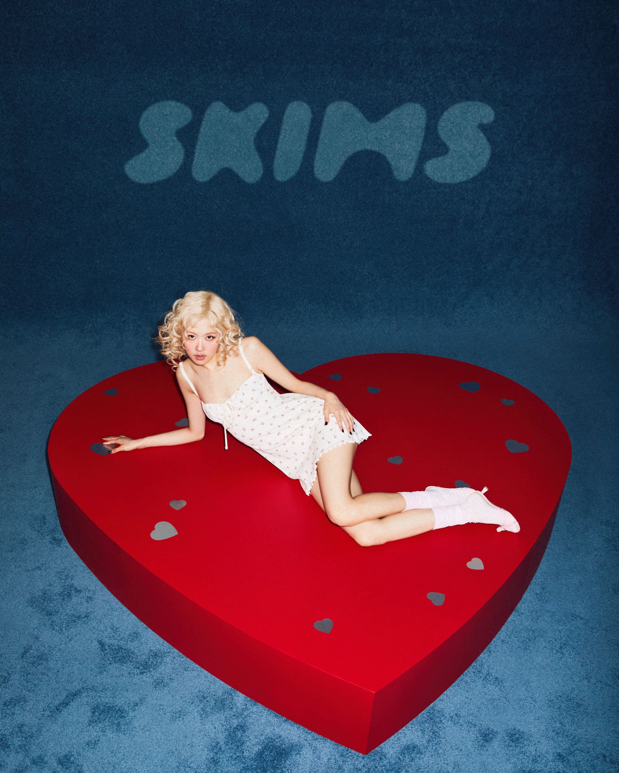 Model with blonde curly hair wearing a white slip dress lying on a large red heart platform against a dark blue textured background.