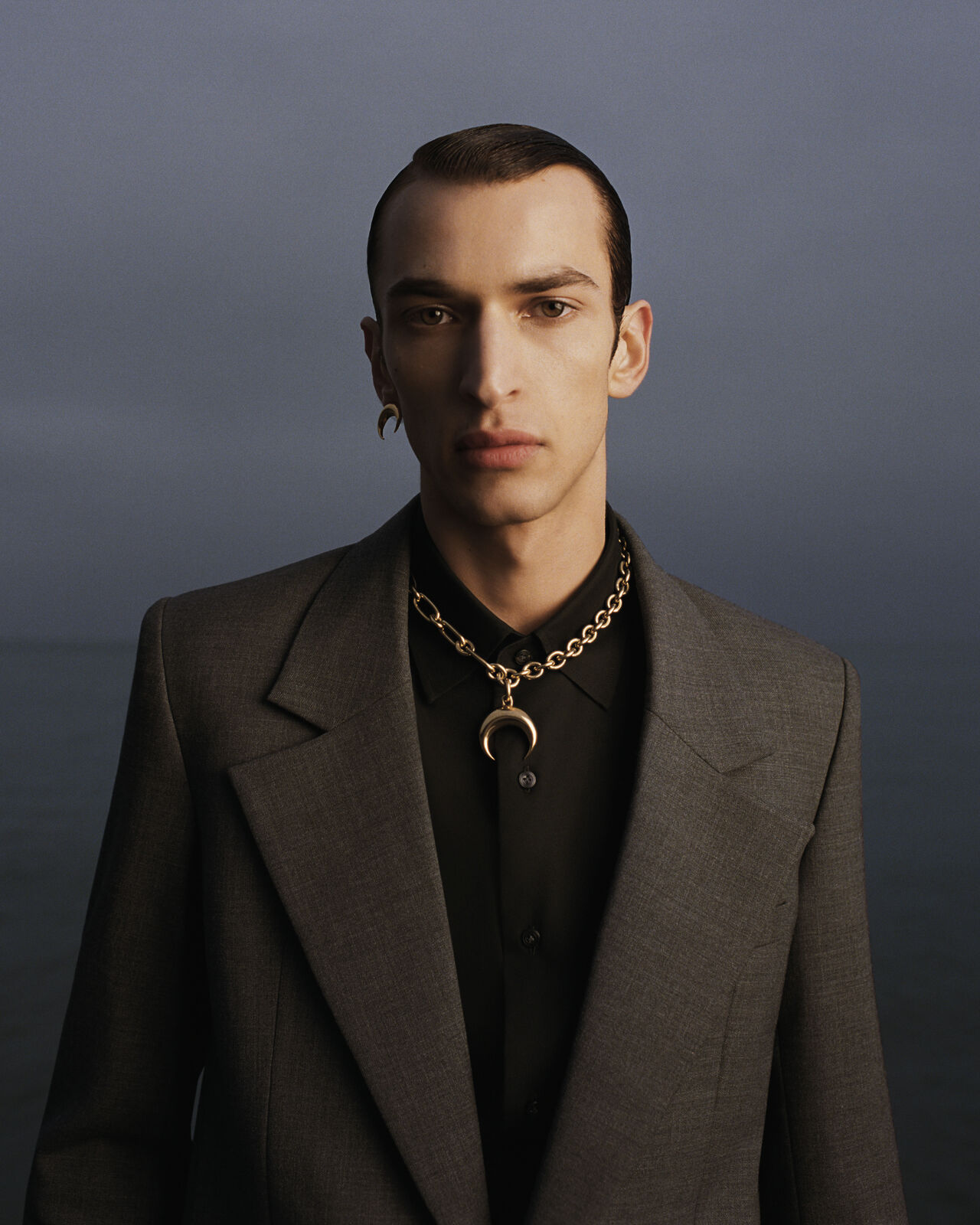Portrait of a man wearing a grey blazer, black shirt, and gold crescent pendant necklace against an overcast sky
