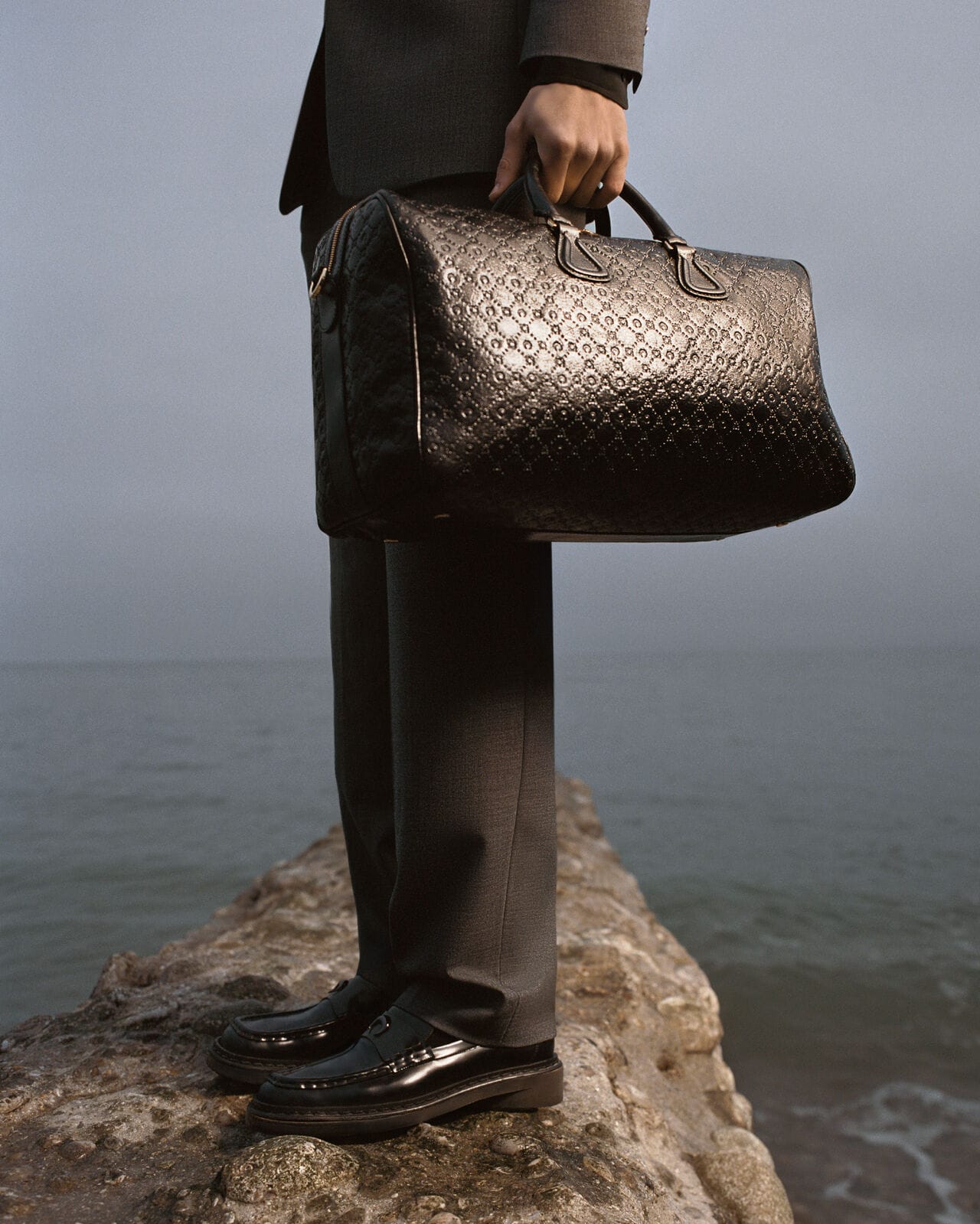 Person holding a large black patterned duffel bag, standing on a rocky coastal ledge by water.
