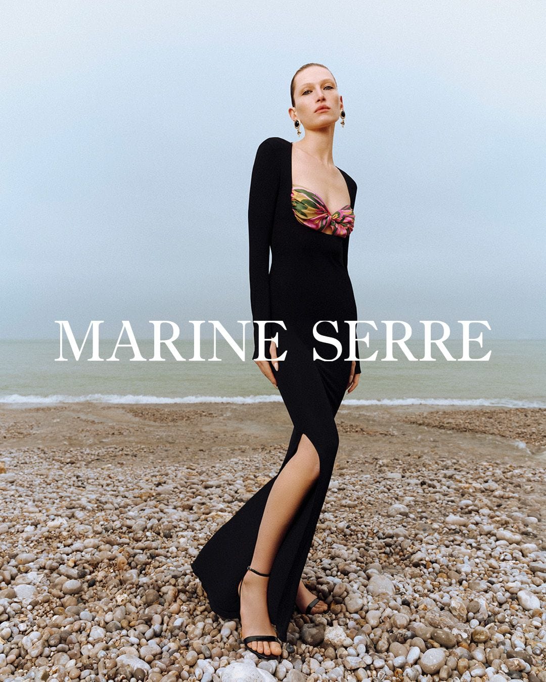 Model standing on a pebble beach wearing a long black dress with a colourful patterned bustier against an overcast sky