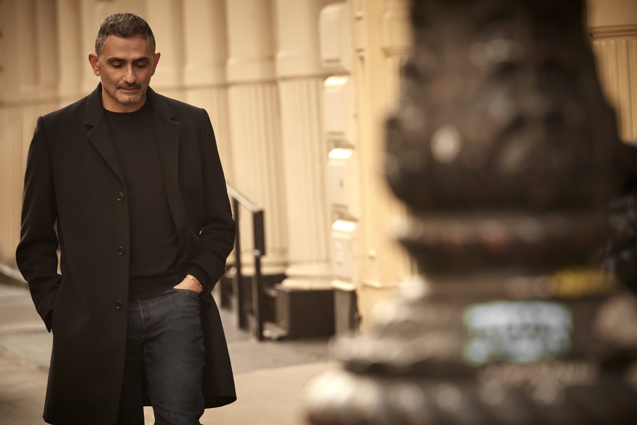 Man wearing a dark coat and blue jeans walking past neoclassical columns on a city street.