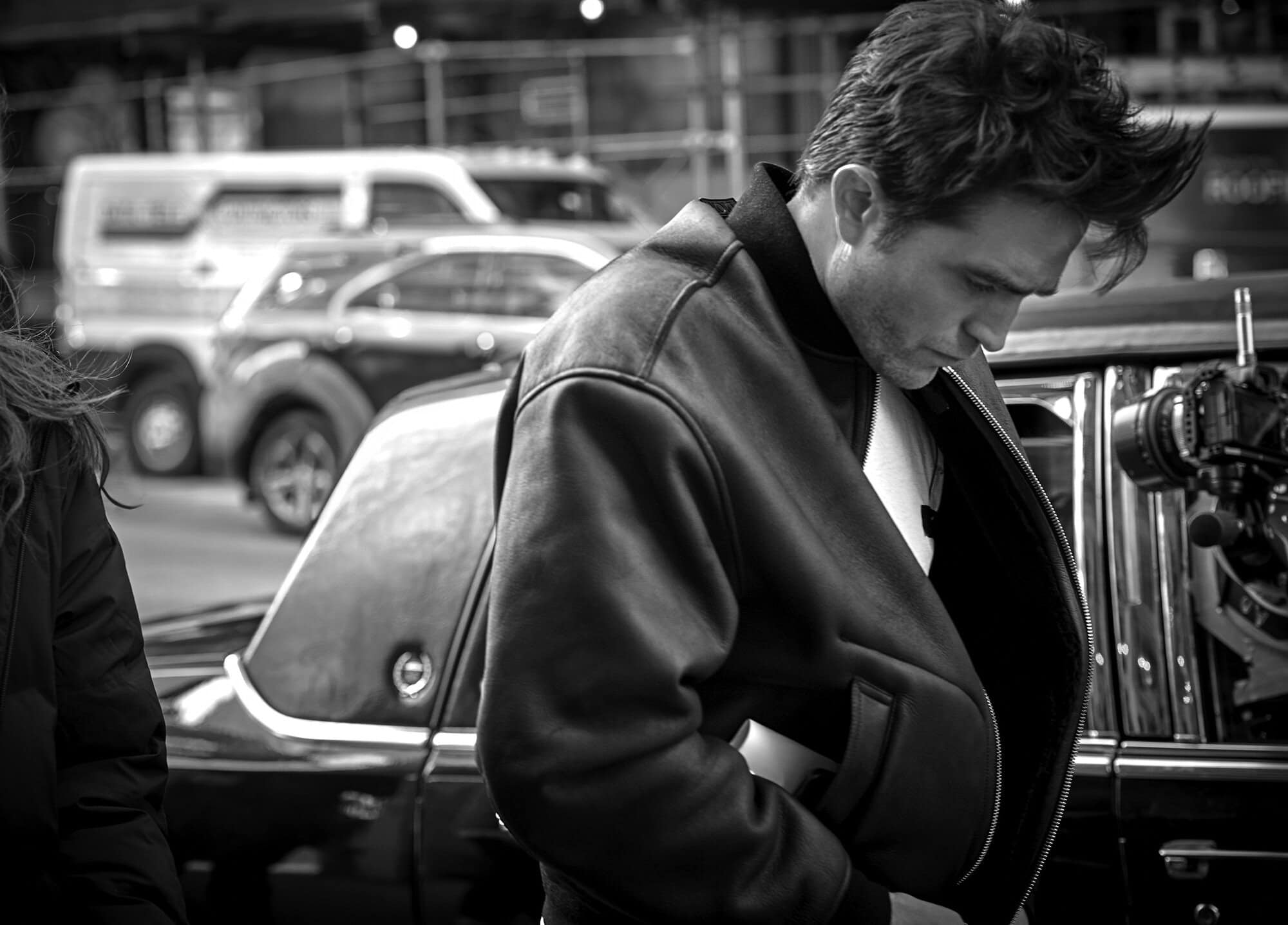 Robert Pattinson wearing a dark leather bomber jacket outdoors near a car for the Dior Homme Parfum ad campaign.