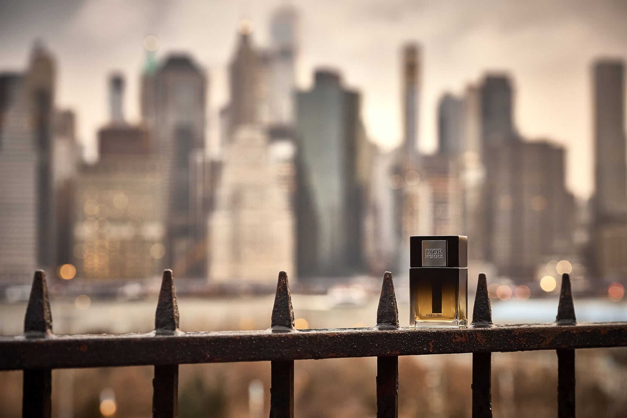 A Dior Homme perfume bottle resting on a dark iron railing overlooking a blurred cityscape skyline at dusk.