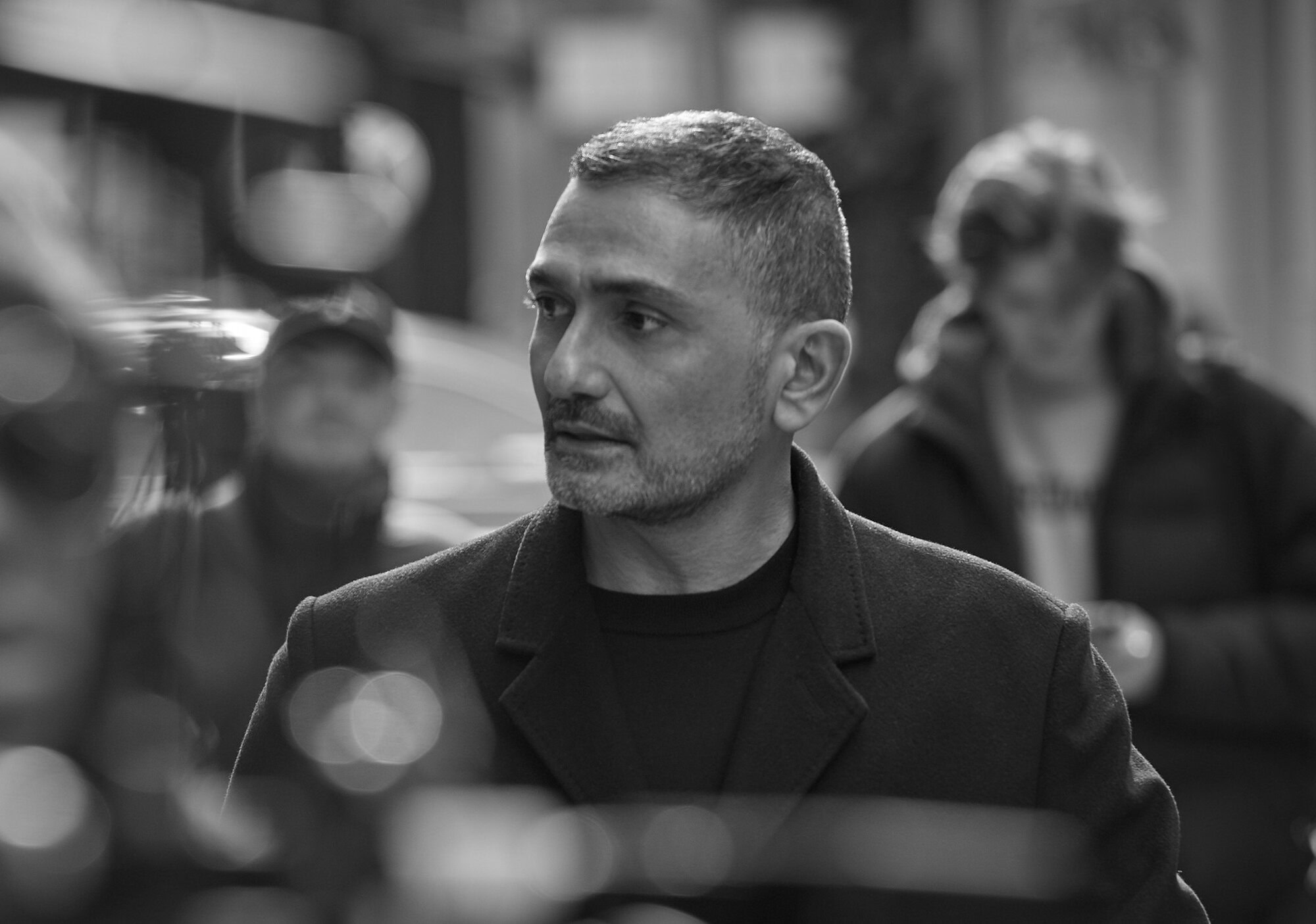 Monochrome portrait of a man in a dark coat and t-shirt outdoors in New York City with blurred figures in the background