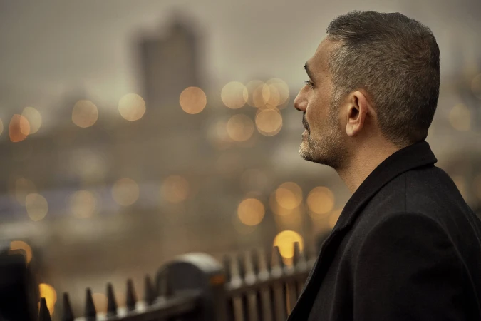 Profile view of a man with short grey hair wearing a dark coat looking out over a city skyline at dusk