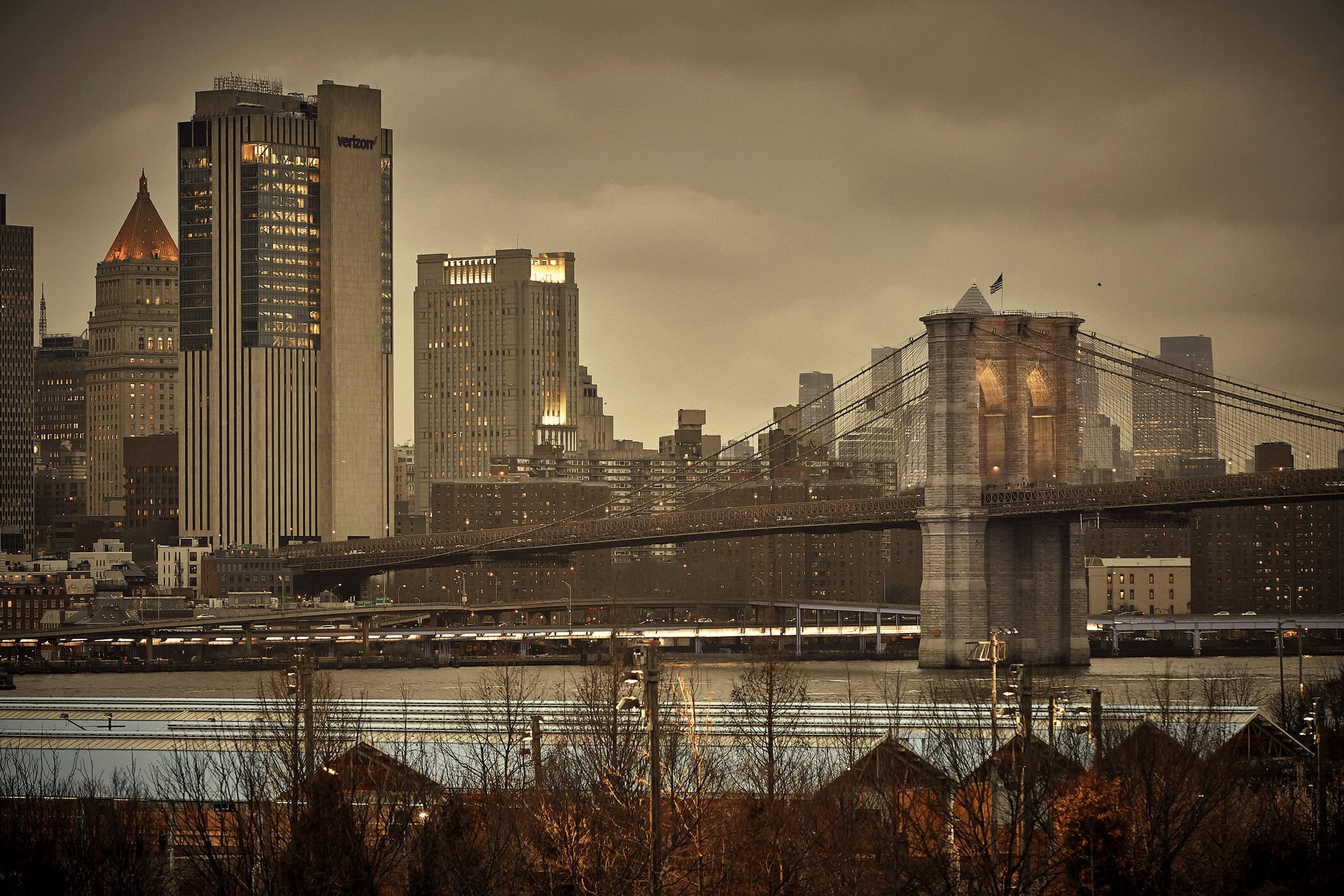 Brooklyn Bridge and downtown Manhattan skyline viewed across the East River under a muted, overcast sky
