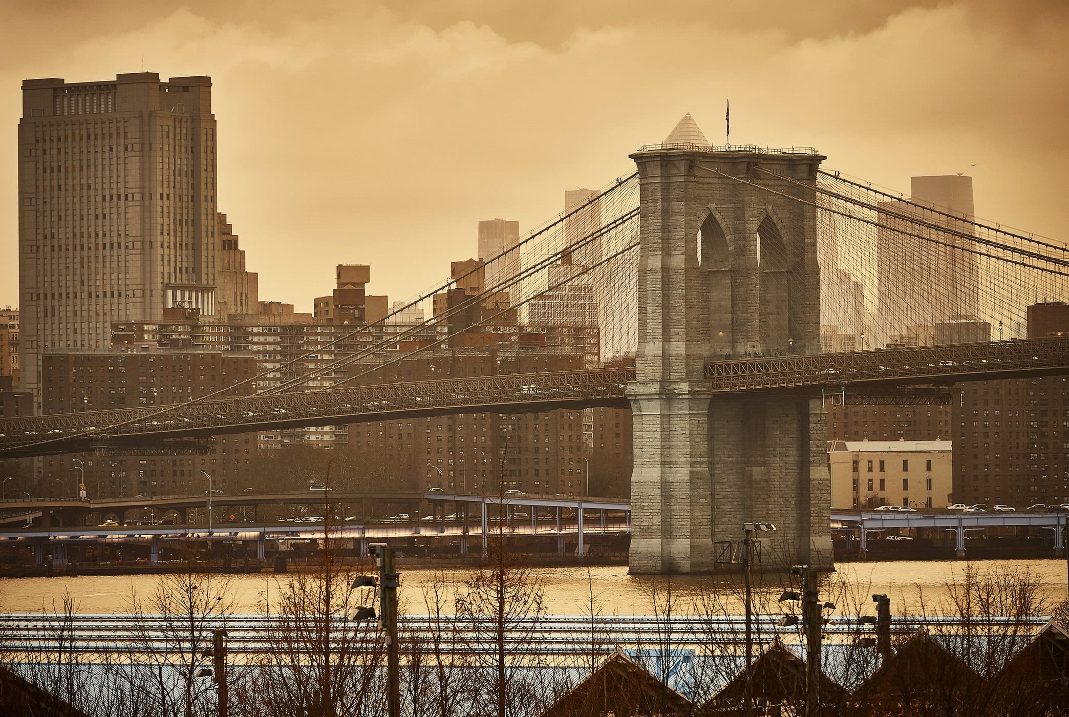 Brooklyn Bridge spanning a wide river with city skyscrapers in the background under a warm sepia sky.