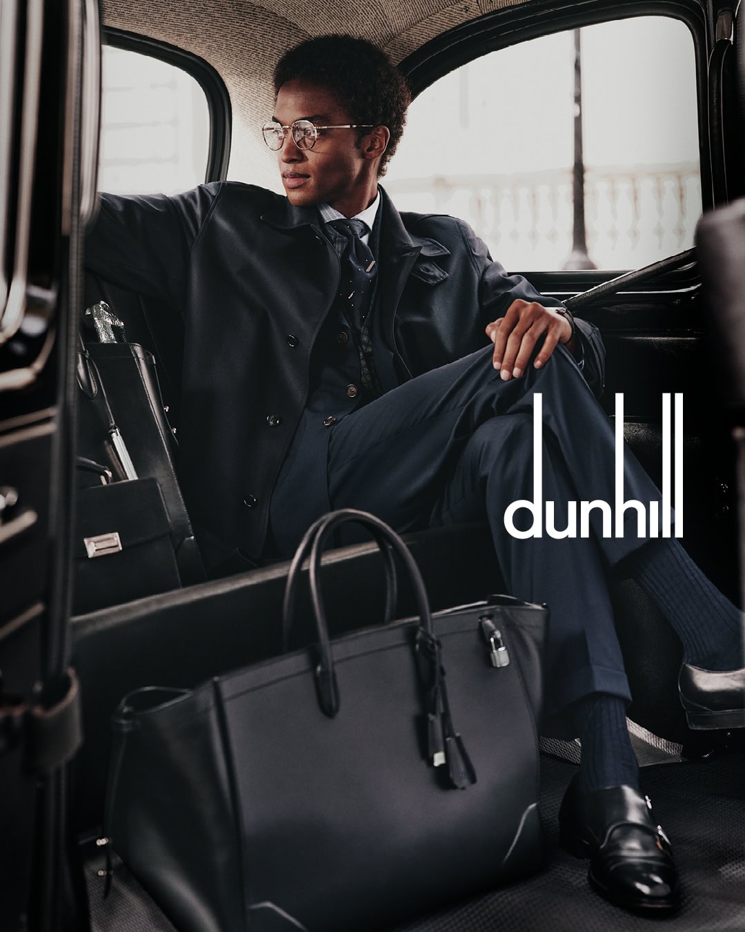 Man wearing a dark suit and glasses seated inside a car with a black leather tote bag on the floor in front of him.