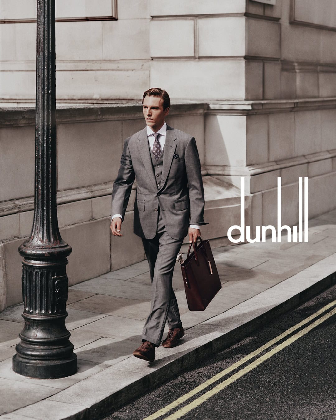 A man in a grey suit holding a leather briefcase walking past a lamppost on a city street for Dunhill.
