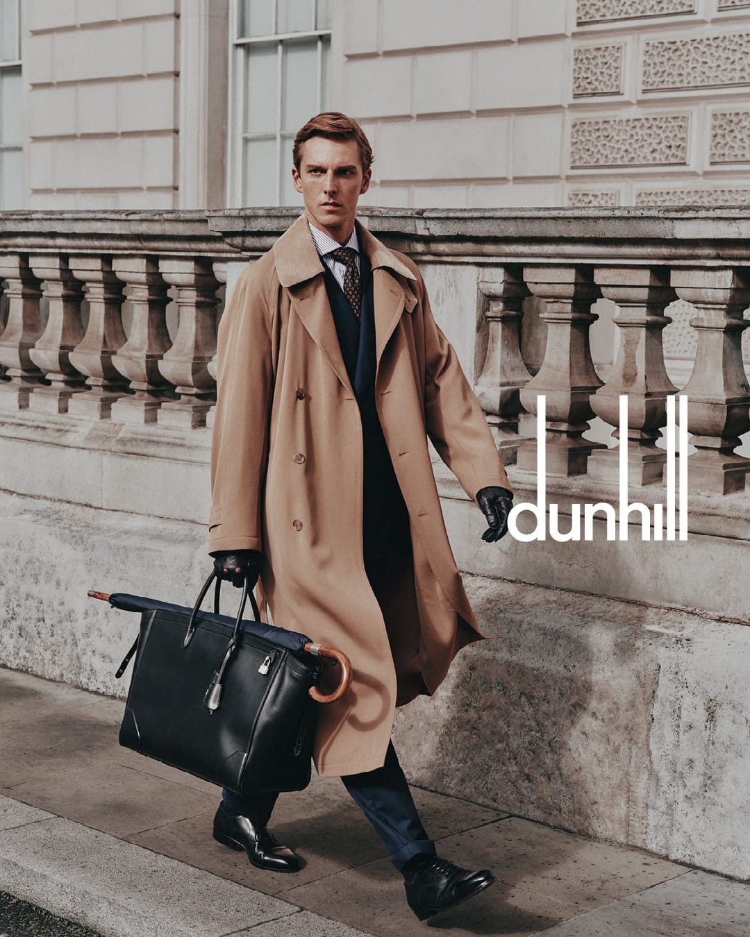 Man in a tan trench coat walking outside while holding a black leather bag and umbrella in front of a stone balcony.