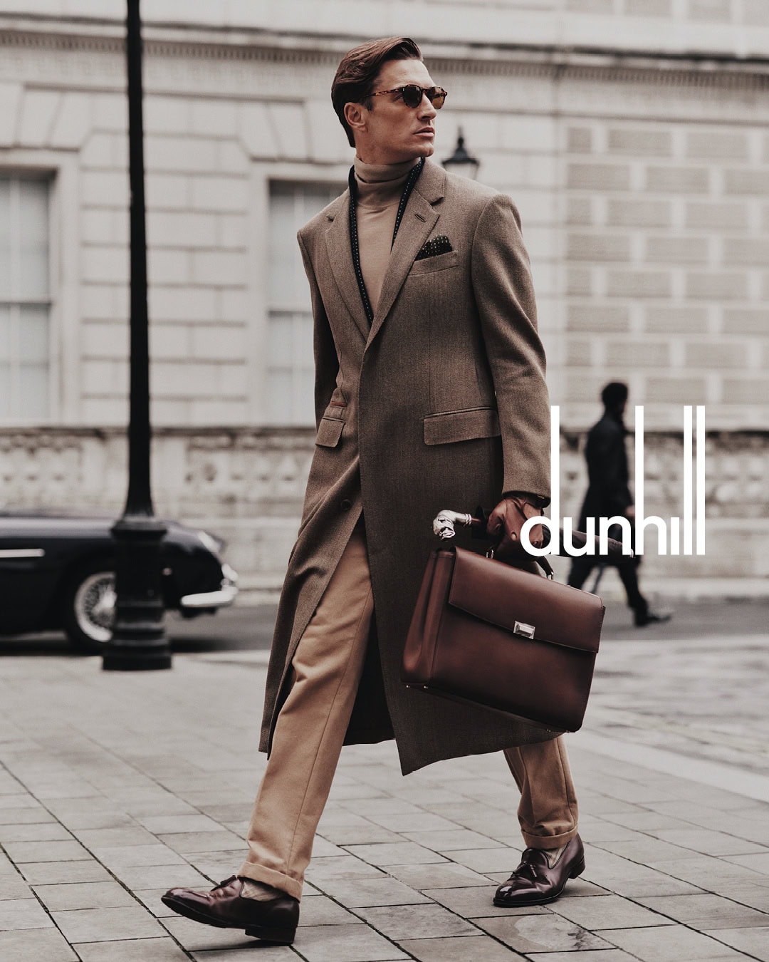 Man wearing a brown overcoat and turtleneck holding a leather briefcase on a paved city street