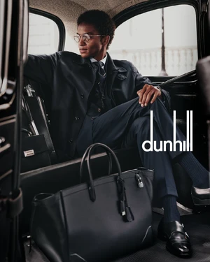 Man wearing a dark suit and glasses seated inside a car with a black leather tote bag on the floor in front of him.