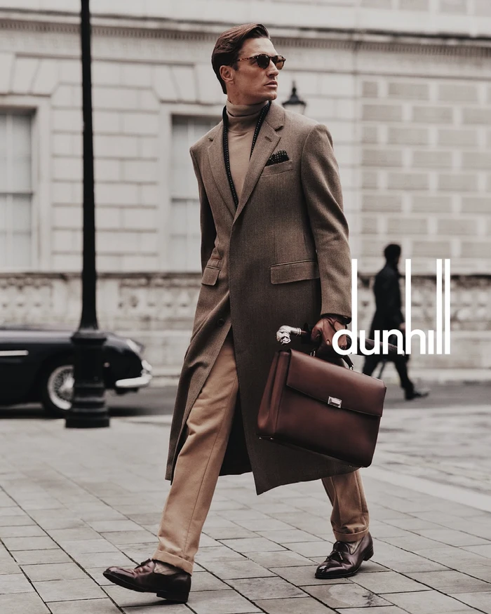 Man wearing a brown overcoat and turtleneck holding a leather briefcase on a paved city street