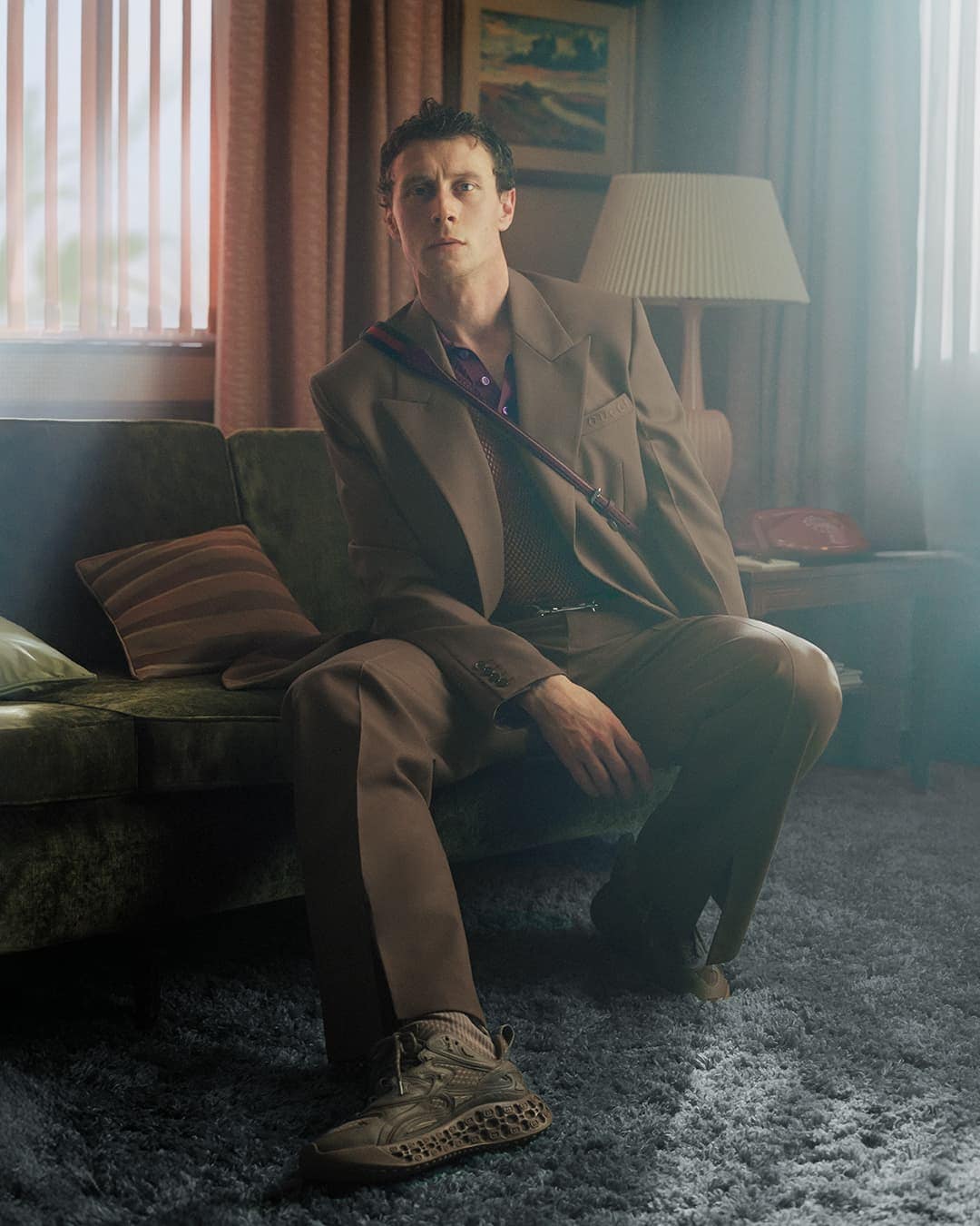 George MacKay seated on a green velvet sofa wearing a brown suit and sneakers in a living room