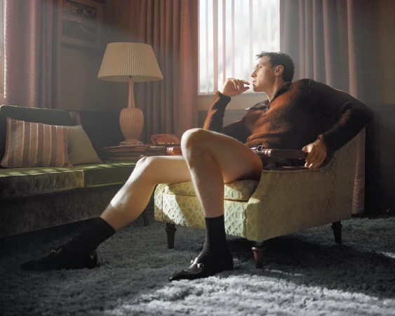Man wearing a brown sweater and black loafers sitting in an armchair inside a living room for Gucci.