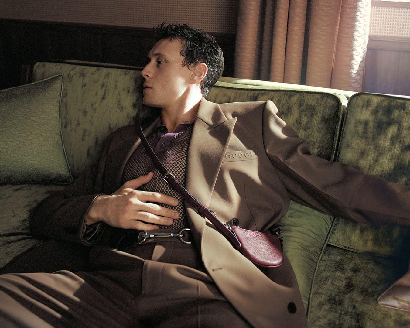 Man wearing a brown suit jacket and mesh polo sitting on a green velvet sofa for Gucci spring 2025 campaign