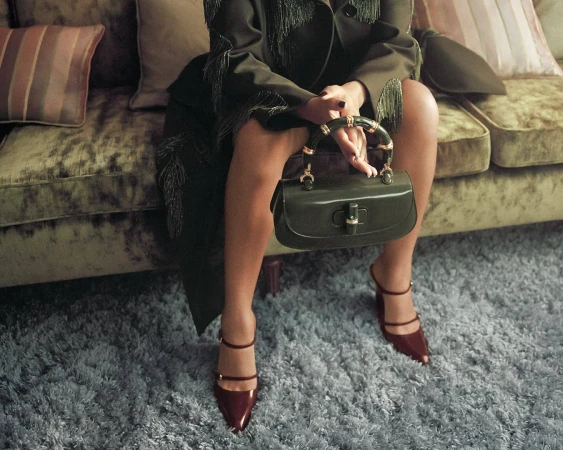 Seated woman holding a glossy green handbag with a bamboo handle wearing a fringe coat and red strappy heels.