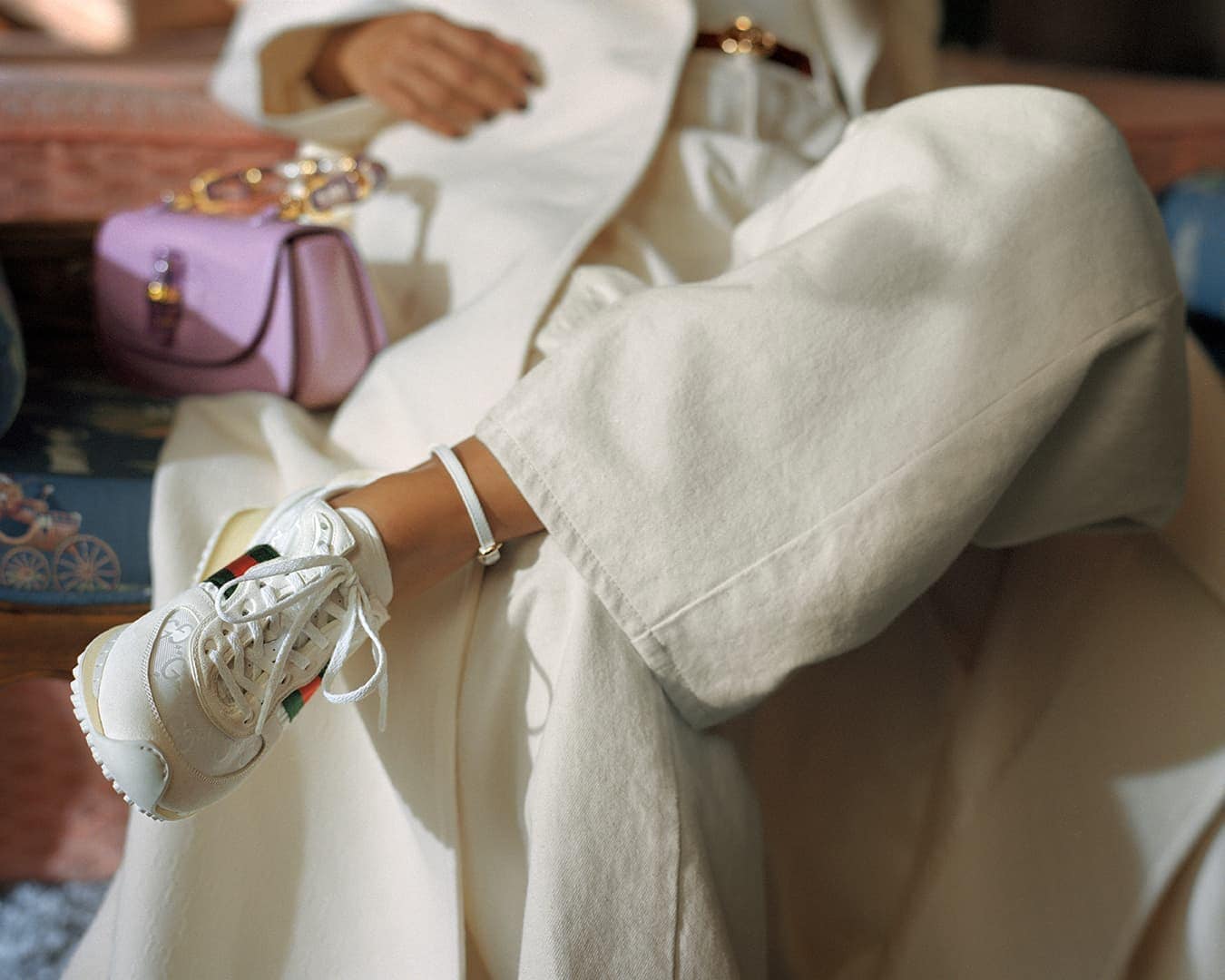 Close-up shot of a person wearing white trousers, white sneakers, and a light-colored coat with a purple handbag resting nearby.