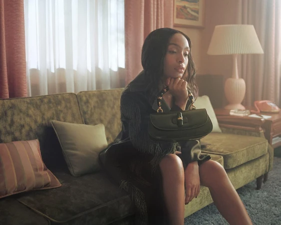 Yara Shahidi seated on a green velvet sofa holding a black handbag inside a retro living room setting.