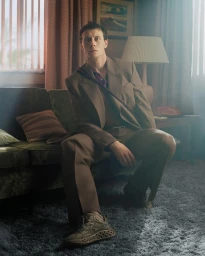 George MacKay seated on a green velvet sofa wearing a brown suit and sneakers in a living room