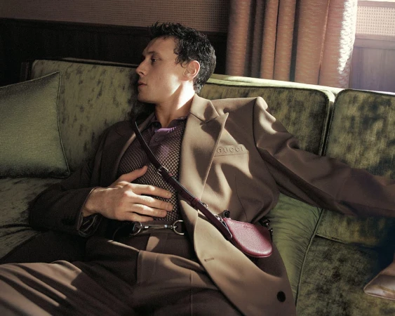 Man wearing a brown suit jacket and mesh polo sitting on a green velvet sofa for Gucci spring 2025 campaign