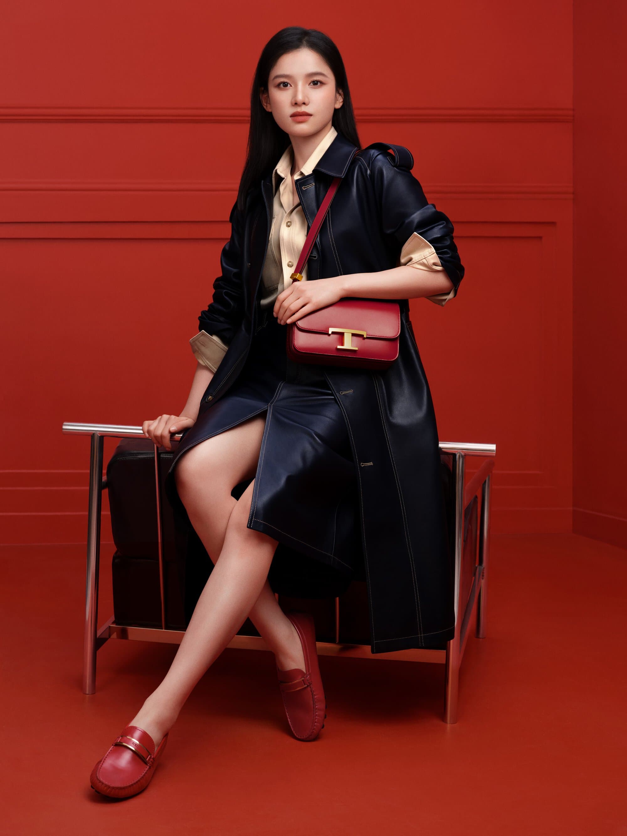 Model Zhang Jinyi seated on a modern frame chair wearing a dark leather coat and red loafers holding a red handbag against a red wall.