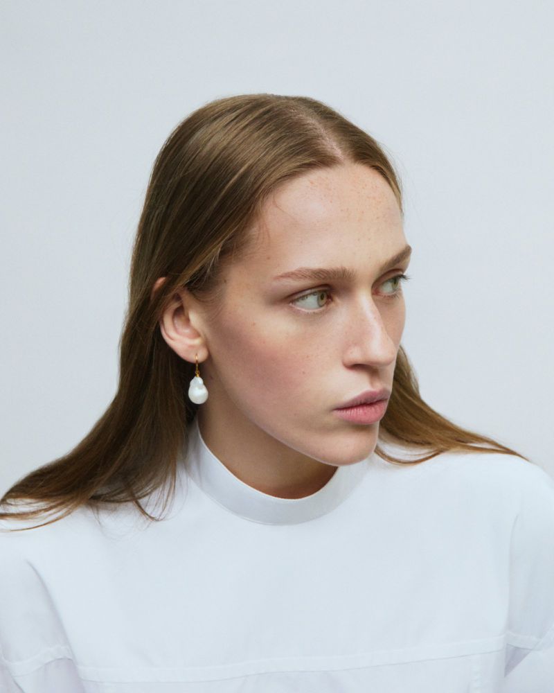 Portrait of an adult female model wearing a white top and a baroque pearl earring, looking away from the camera in a neutral studio setting.