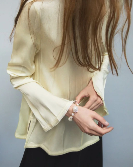 Model adjusts a pearl and gemstone bracelet on her wrist, wearing a pale yellow blouse with long sleeves against a light gray background.
