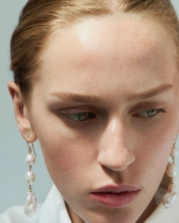 Close-up portrait of a light-skinned woman with an understated expression, wearing pearl drop earrings against a soft, neutral background.