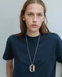 Close-up portrait of a woman wearing a navy blue t-shirt and a long silver pendant necklace against a plain grey studio background.