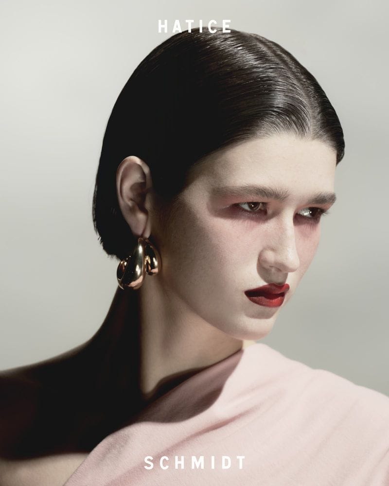 Close-up portrait of a model with dark slicked-back hair, bold red lipstick, and gold earrings wearing a light pink top against a grey background