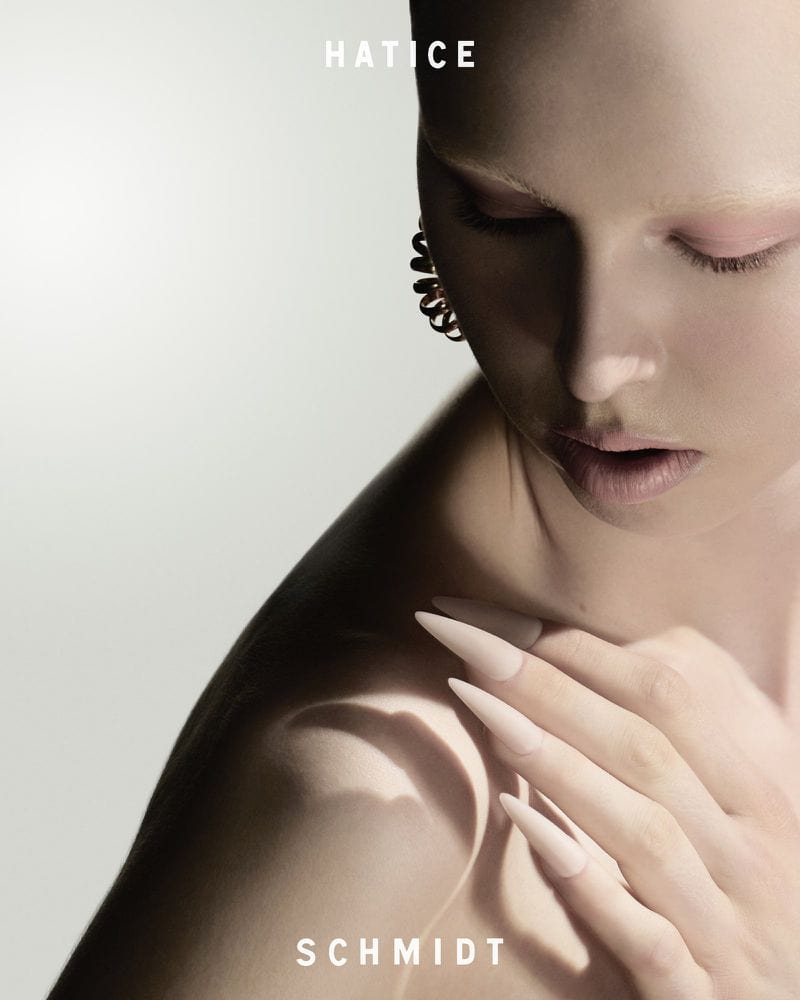 Close-up portrait of a model with long pointed nails resting on her shoulder against a neutral grey seamless background