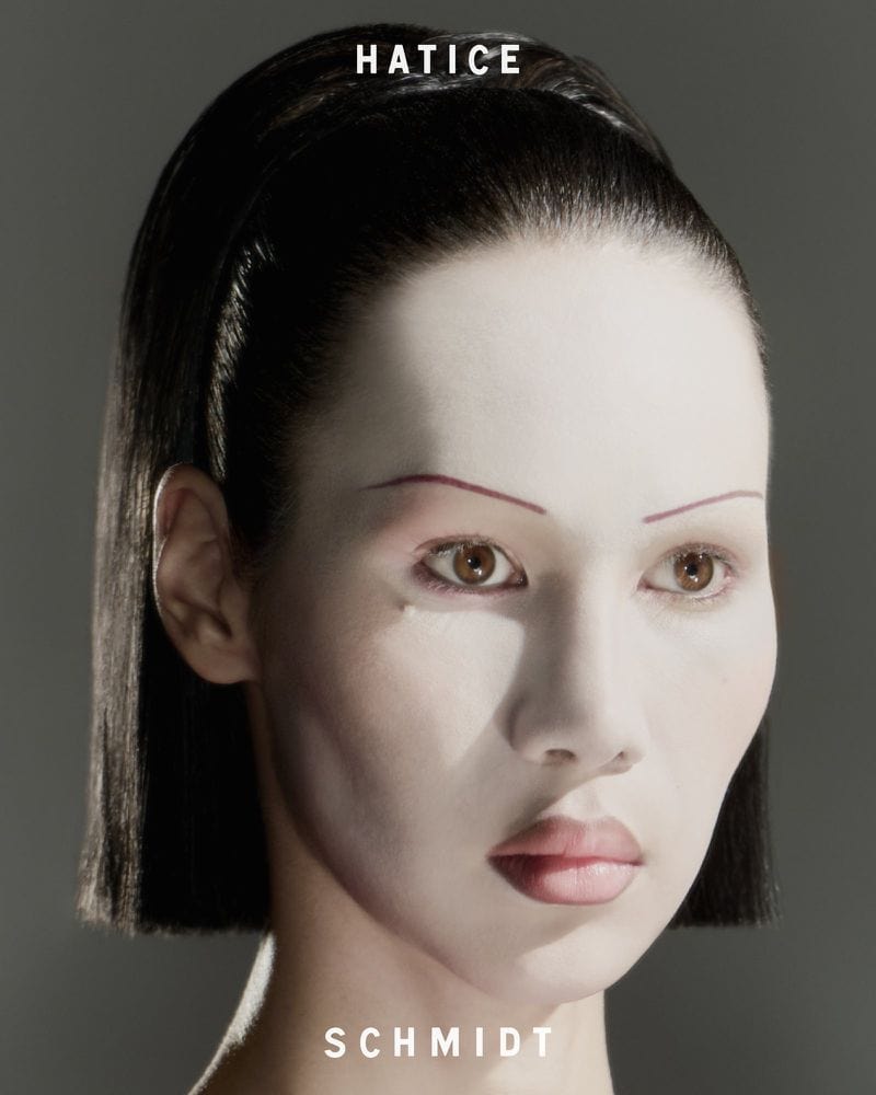 Close-up portrait of a woman with dark hair slicked back and pale makeup against a grey background for a beauty campaign.