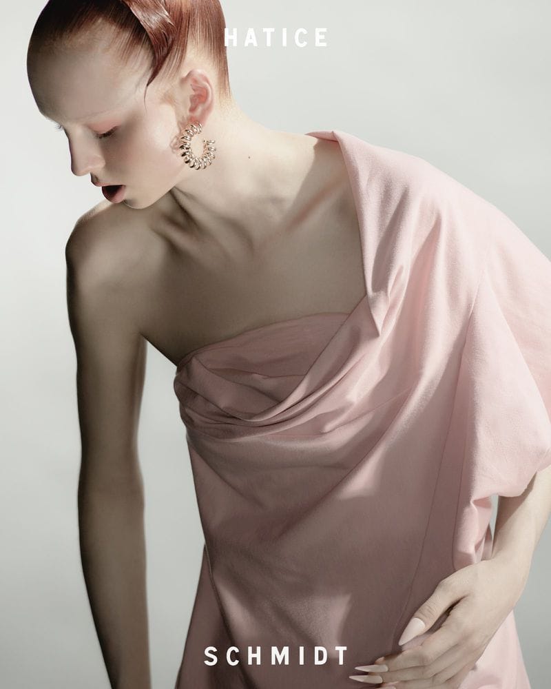 Model in pale pink dress with sculpted hair and gold hoop earrings against a neutral grey background